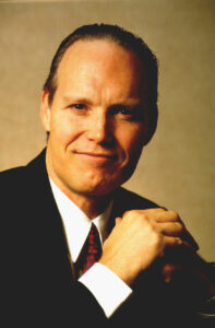 Patrick Reynolds sits smiling in a classic male power pose in a suit with burgundy tie, hair hair slicked back.