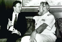 Patrick in black suit, sits on a couch engaged in conversation with Surgeon General Koop in black and white photo.