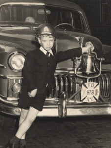 Patrick Reynolds, wearing a fireman's hat, stands in front of a fire truck with a tag FD 878 NY.