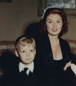 A very young Patrick sits beside his mother wearing a suit with black tie.