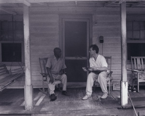 Patrick sits in an all white outfit speaking to a man on the front porch with swing.