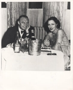 Dick and third wife, Muriel Greenough Reynolds sit at a table smiling with a bottle of champagne.