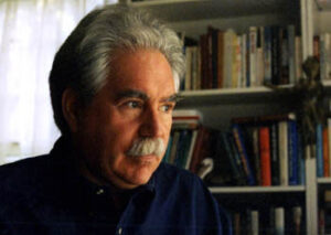 Tom Shachtman sits with sunlit face in a home library wearing a dark navy shirt with gray hair and a gray mustache, gazing out a window. 